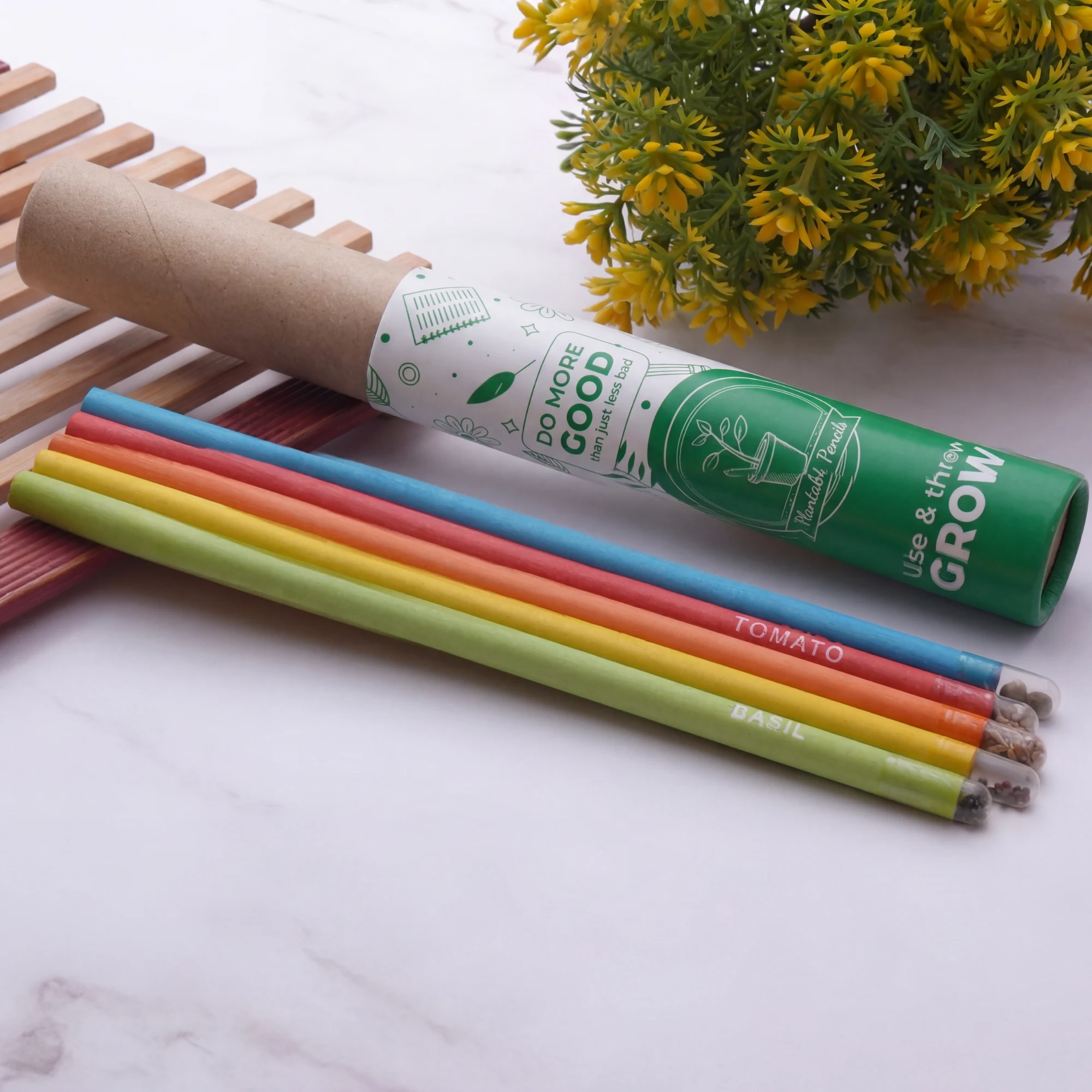 A kraft tube wearing the standard bioQ wrap, printed do more good than just less bad and use and then grow, lying open beside five coloured pencils named tomato and basil, each with a clear seed capsule at its end